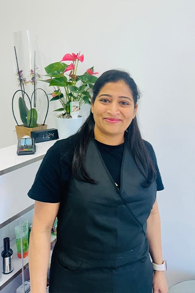 A woman with dark hair is standing indoors, smiling at the camera. She is wearing a black apron over a black shirt. Behind her is a white wall with a shelf displaying decorative plants and various items.