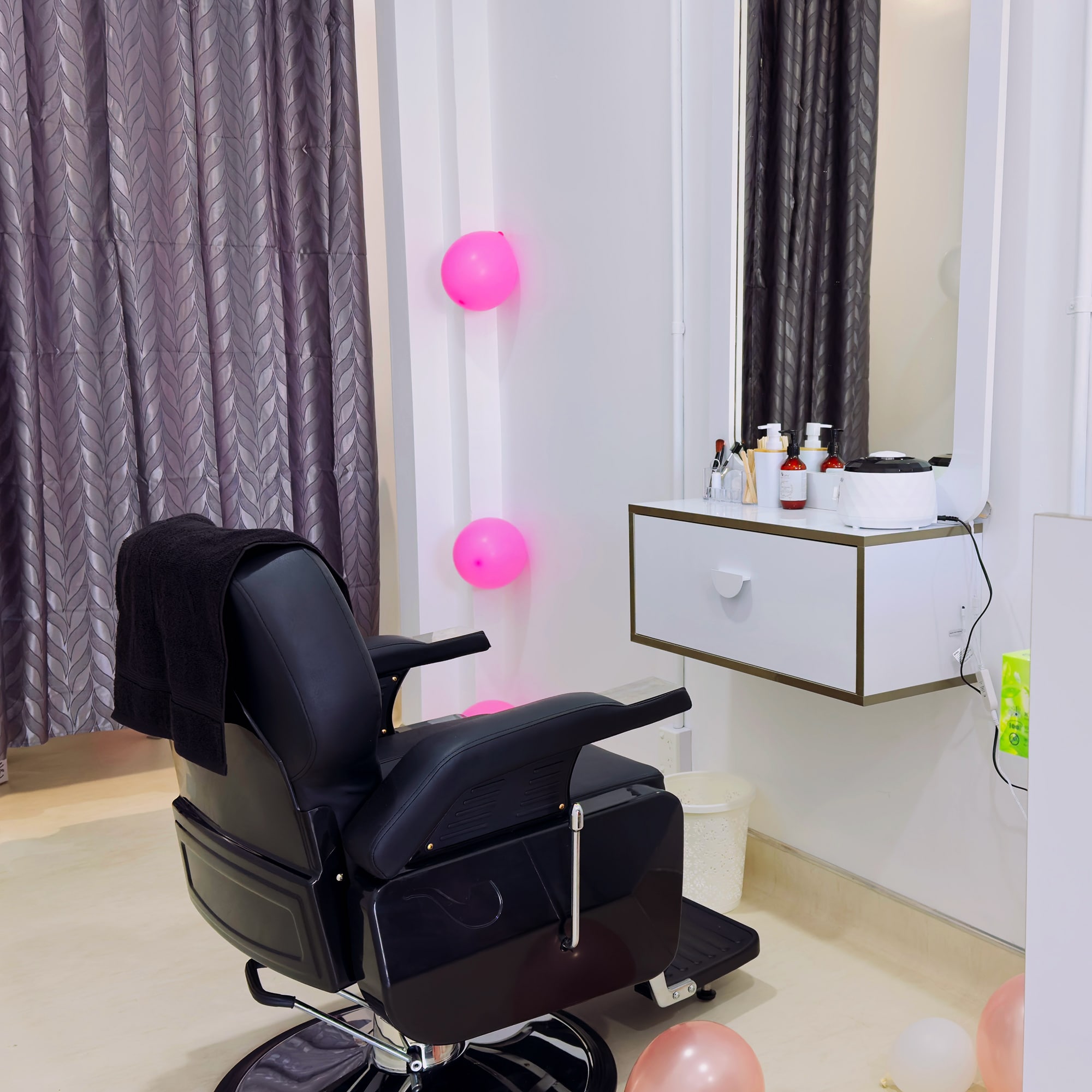 A modern salon chair is positioned in front of a mirror with a white counter containing various hairstyling products. The room has light-colored walls and floor, gray curtains, and pink and peach balloons scattered about.