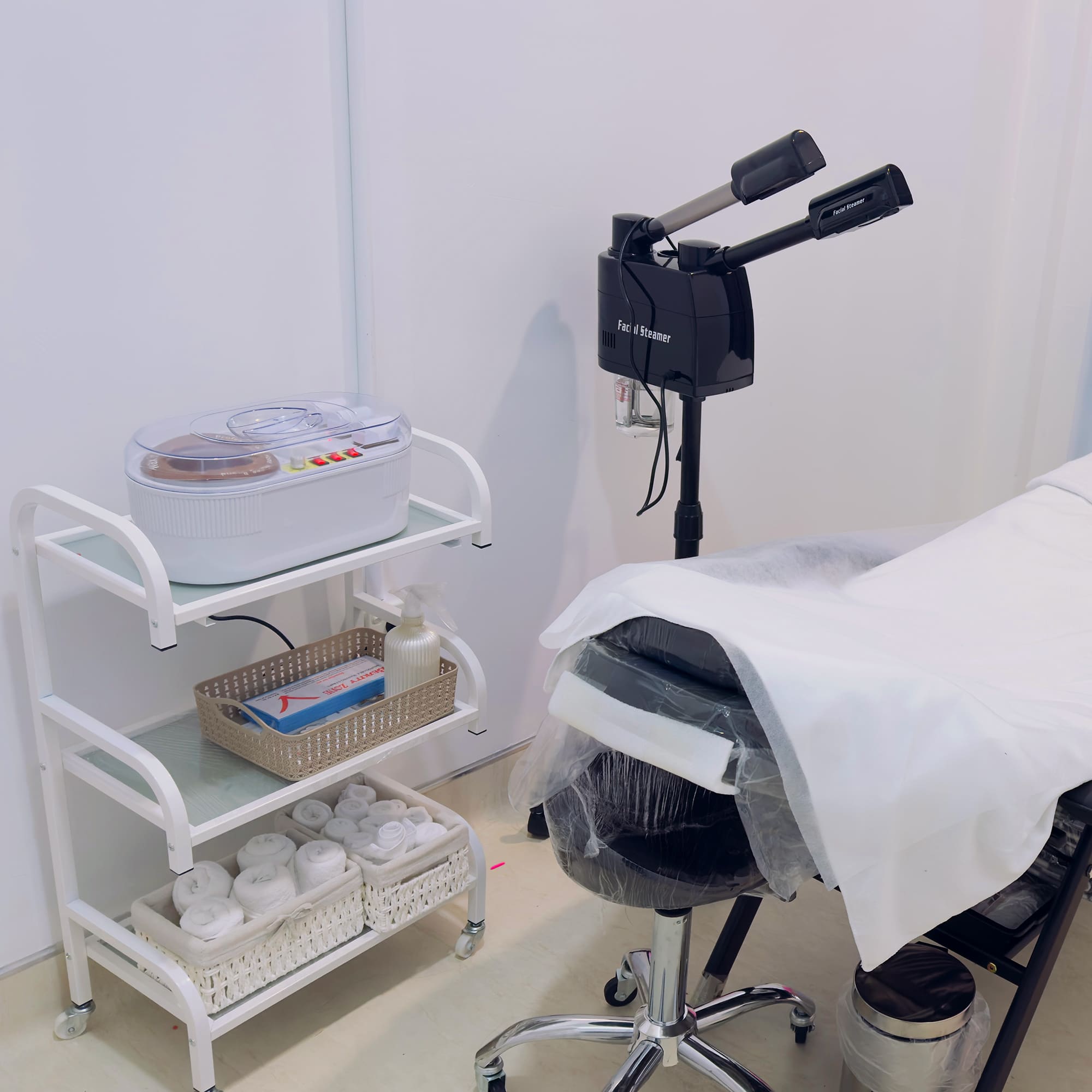 A professional skincare treatment room featuring a white cushioned chair covered with tissue paper, a mobile equipment rack with skincare supplies, a magnifying lamp, a facial steamer, and clean, minimalist walls.