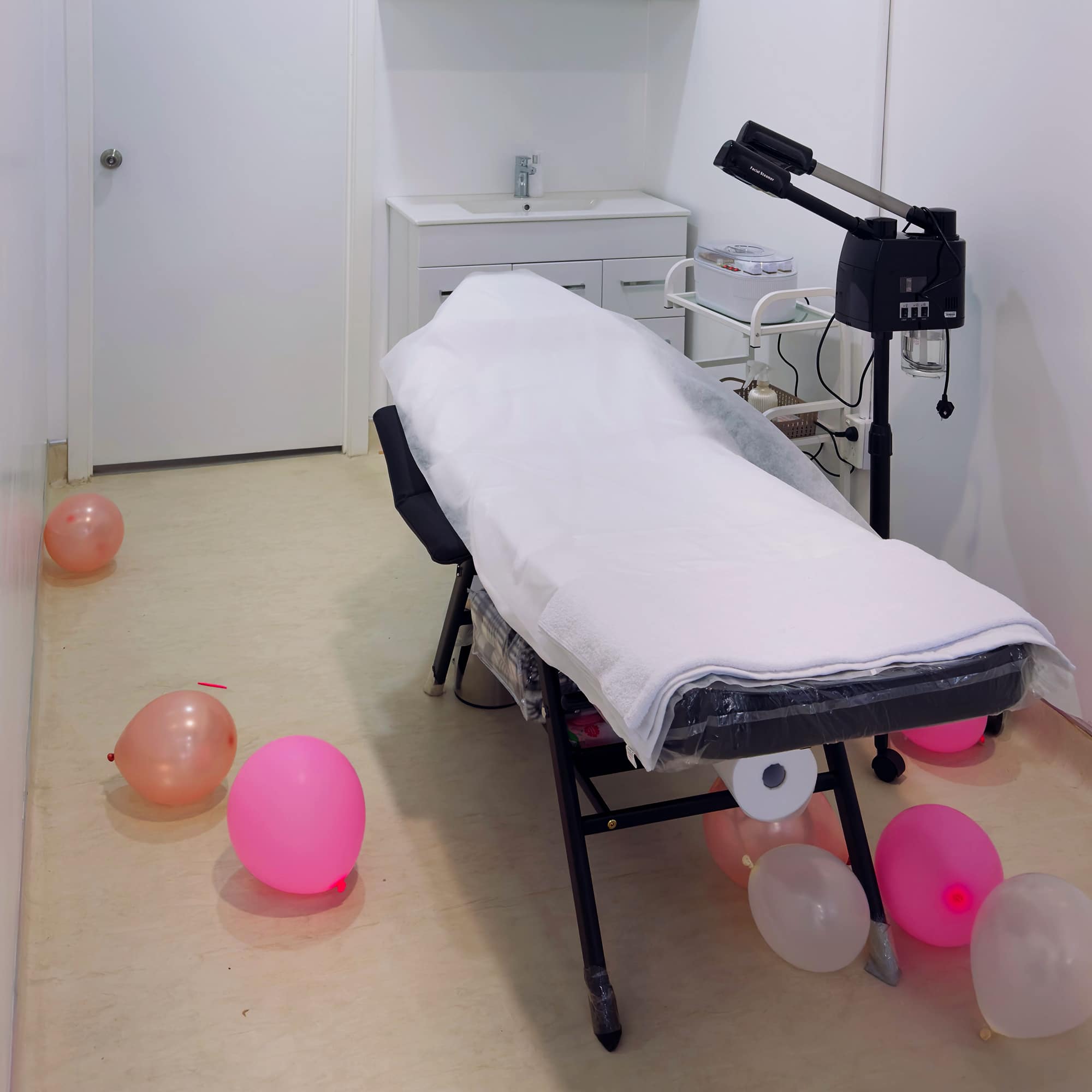 A small, white-walled room with a medical examination table covered in white sheets. Various colored balloons are scattered on the floor around the table. Medical equipment is present, including a sink, shelves, and a rolling cart with instruments.