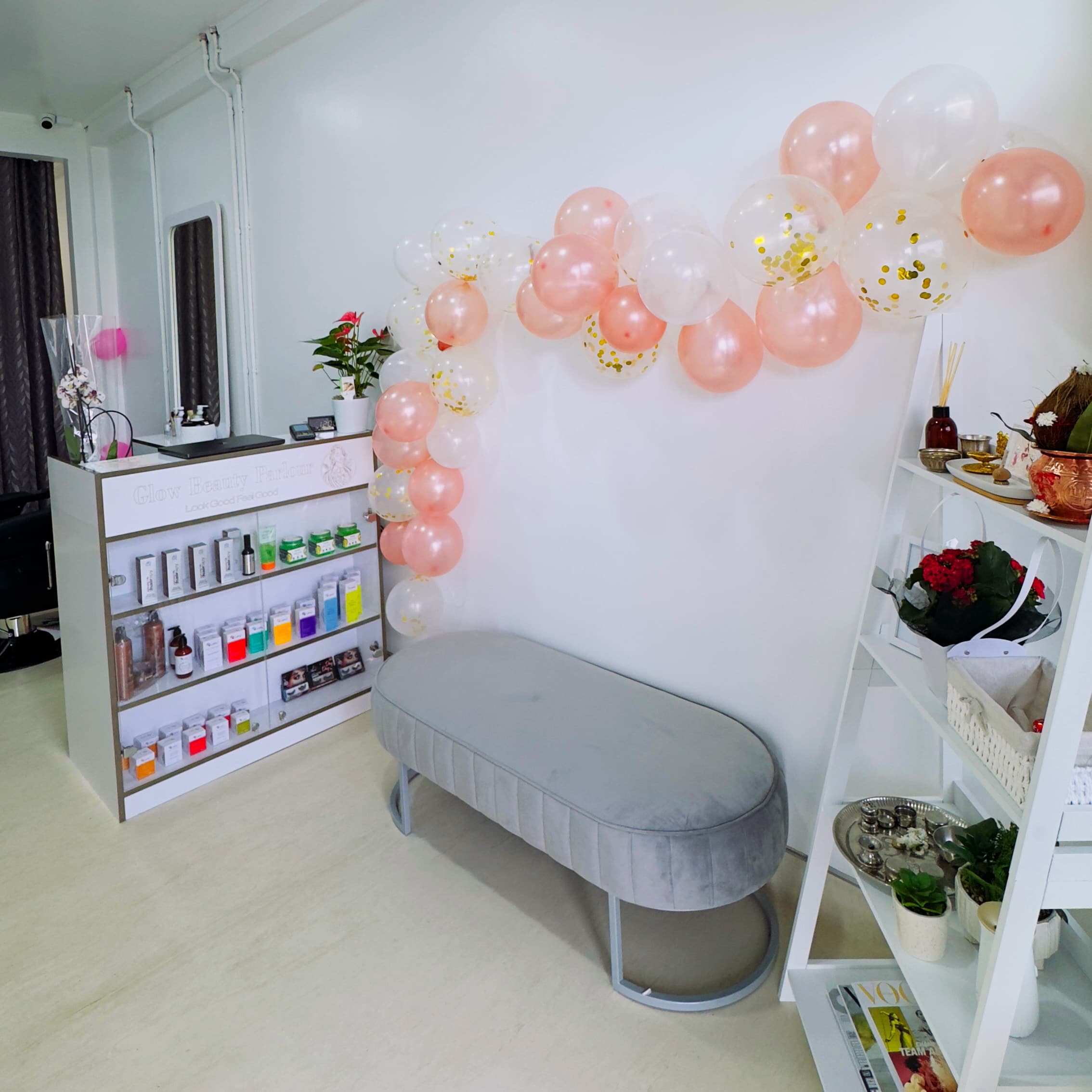 A cozy beauty salon features a wall adorned with pink and white balloons. Beneath the balloons is a stylish grey bench. Shelves on the left hold various beauty products, while a white ladder shelf on the right displays decorative plants and magazines.