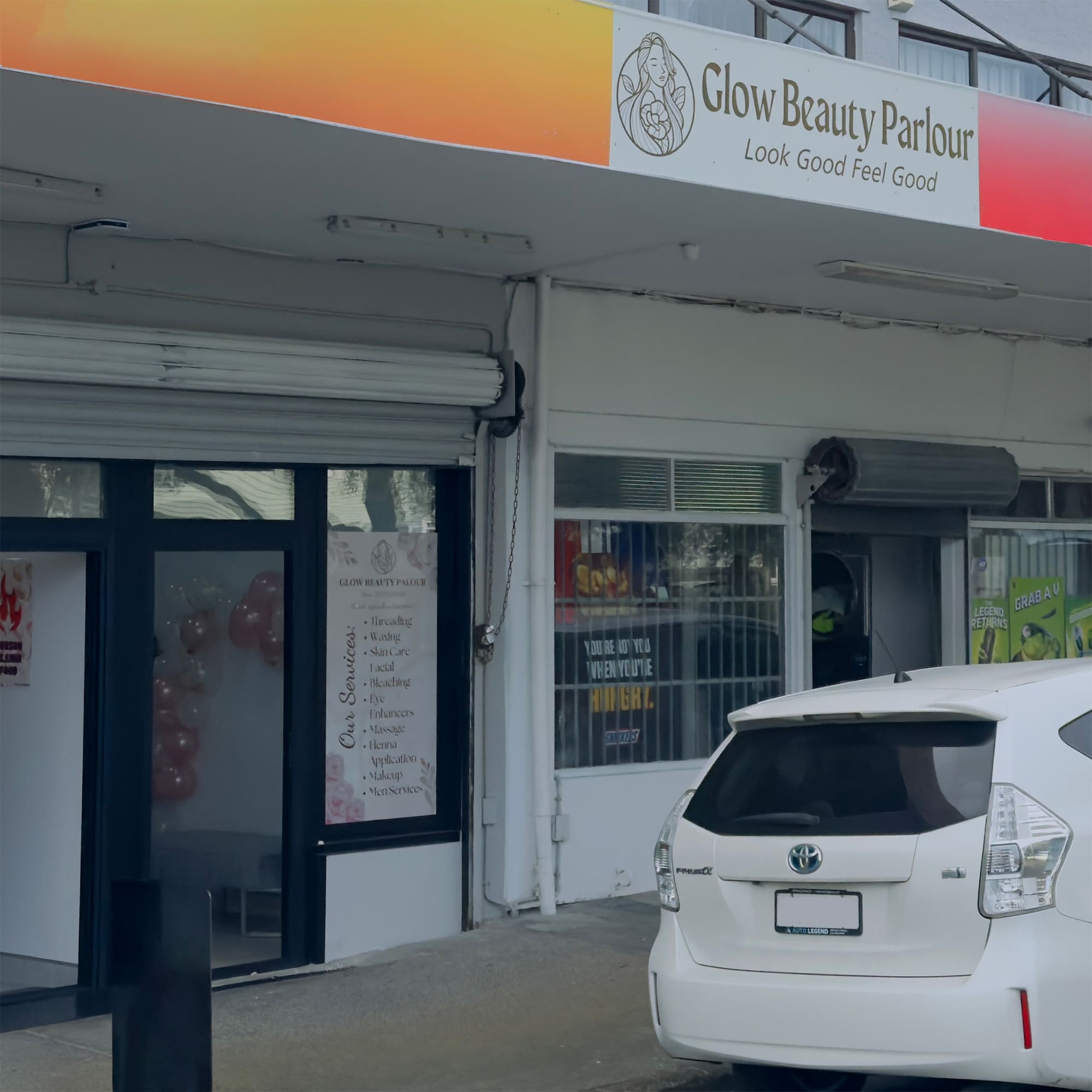 A street view of Glow Beauty Parlour with a sign that reads "Glow Beauty Parlour: Look Good Feel Good." A white car is parked in front. The building has roll-up doors with one partially open, revealing pink balloons inside the parlour. Another shop is visible next door.