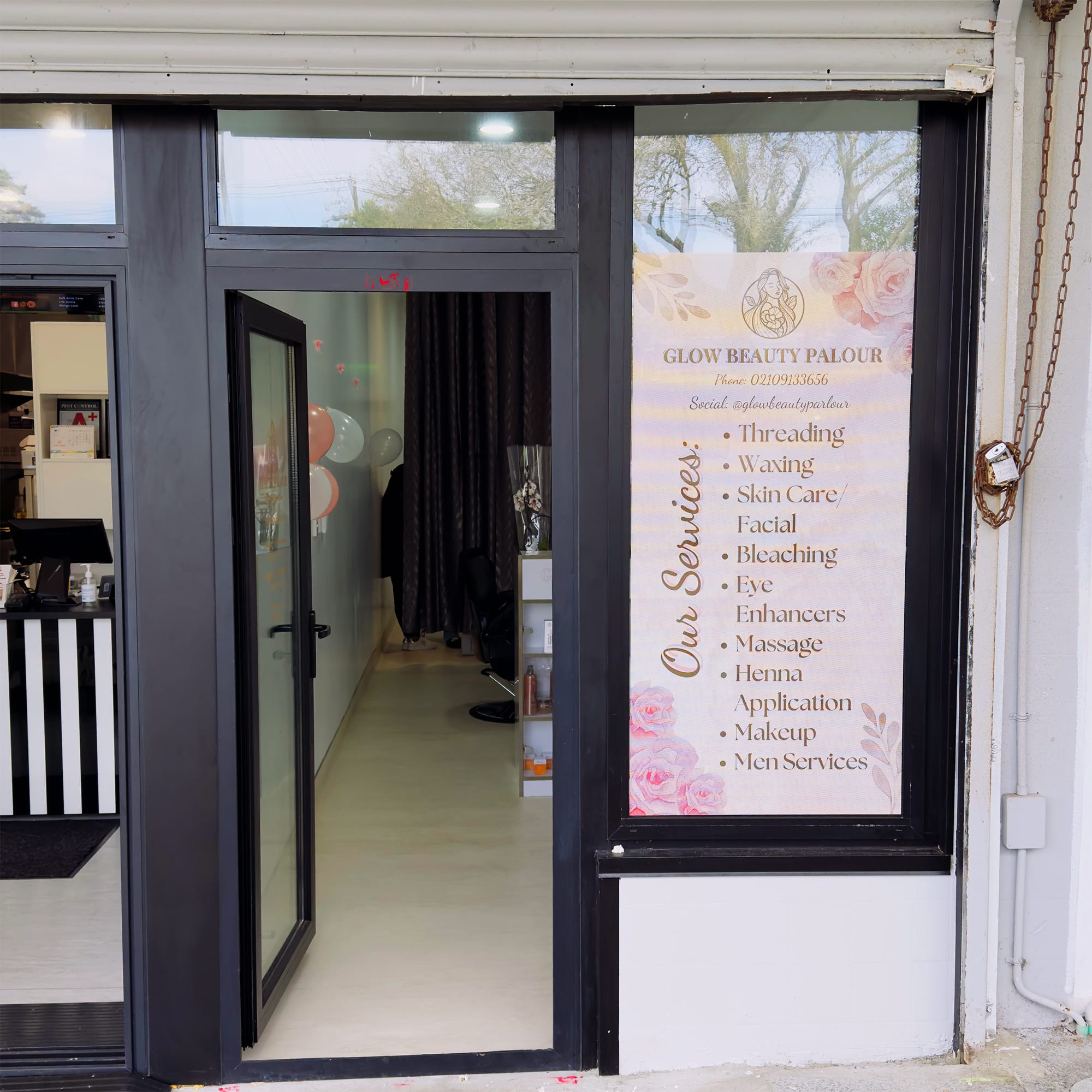 A beauty parlor entrance with a glass door and window. Beside the door, a sign lists services: threading, waxing, skin care, facial, bleaching, eye enhancers, massage, henna, application, makeup, men services. The interior is partially visible through the door.
