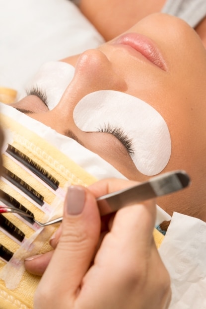At a Beauty Parlour in Auckland, a person is receiving an eyelash extension treatment. They lie peacefully with closed eyes, adhesive pads beneath them. Expert hands gently apply lashes using tweezers and a brush, ensuring a flawless finish.